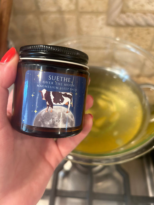 Hand holding 4oz amber glass jar of Suethe Over the Moon Magnesium Sleep Balm with cow illustration and moon label in front of melted balm on stovetop.